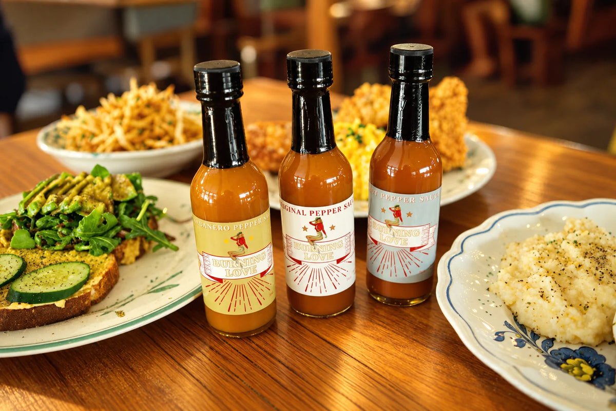 Lady May pepper sauces surrounded by plated restaurant dishes on a wooden table