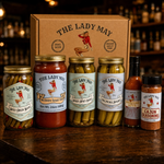 The Lady May Deluxe Bloody Mary Kit lineup on a rustic wood bar – Bloody Mary mix, pickled okra, dill pickles, green beans, hot sauce and Cajun seasoning