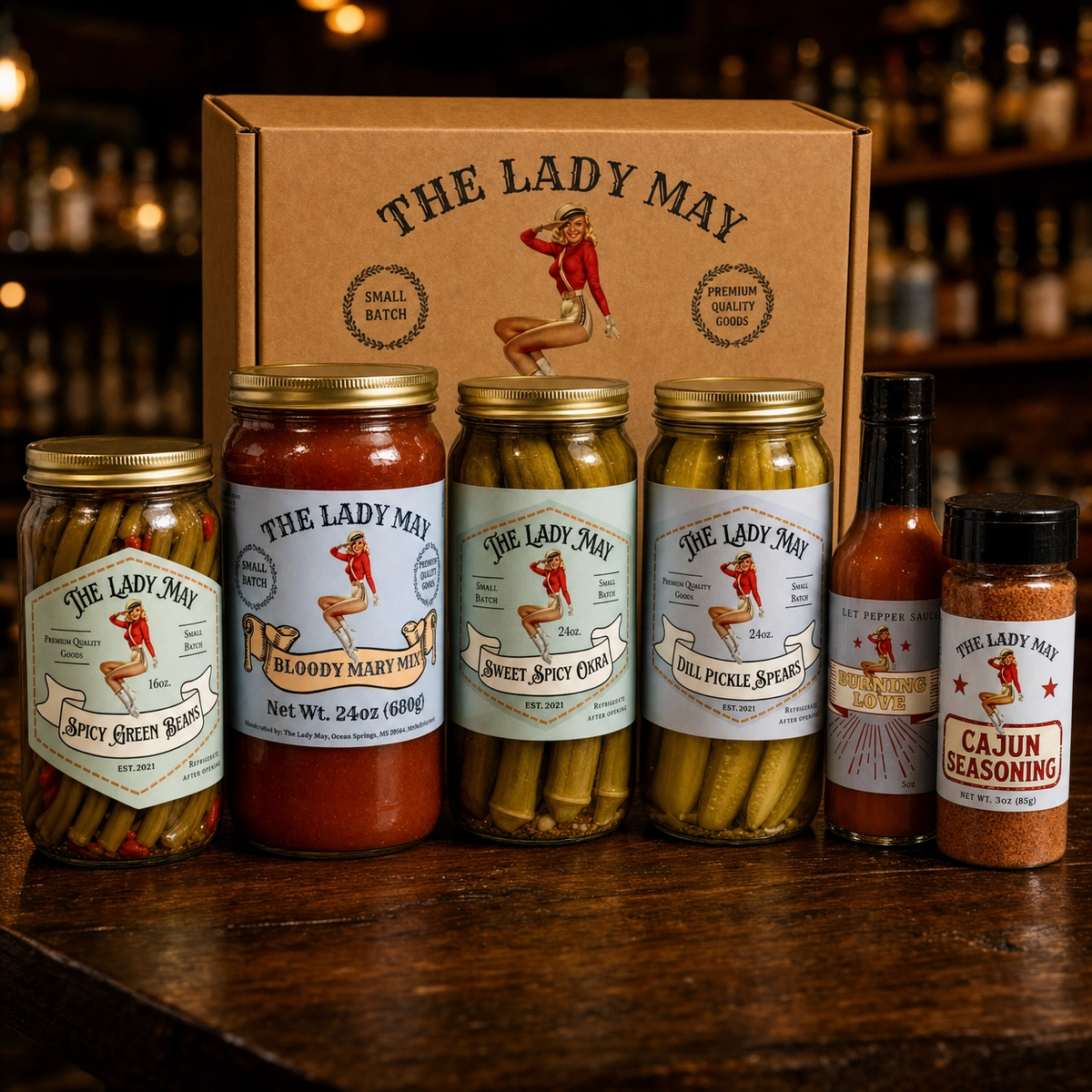 The Lady May Deluxe Bloody Mary Kit lineup on a rustic wood bar – Bloody Mary mix, pickled okra, dill pickles, green beans, hot sauce and Cajun seasoning